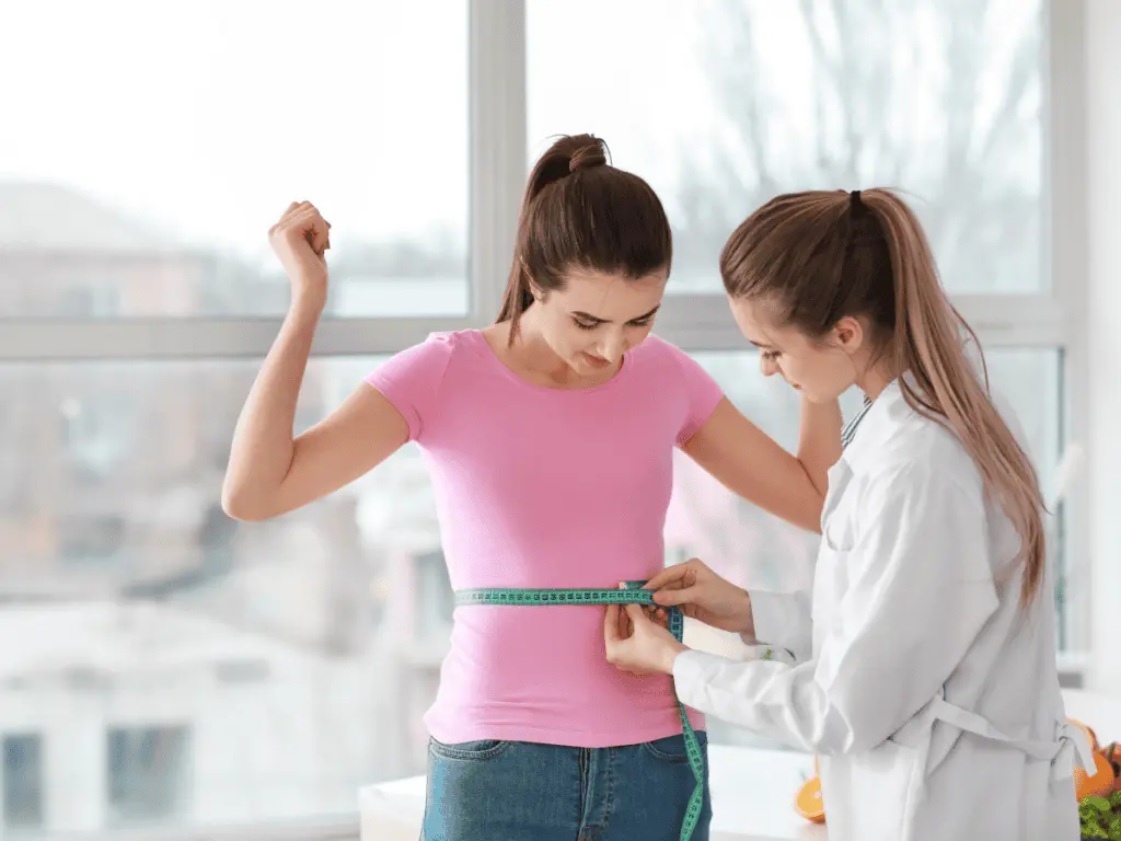 A healthcare professional measures a young woman's waist with a measuring tape during a wellness or weight loss consultation in a bright medical office.