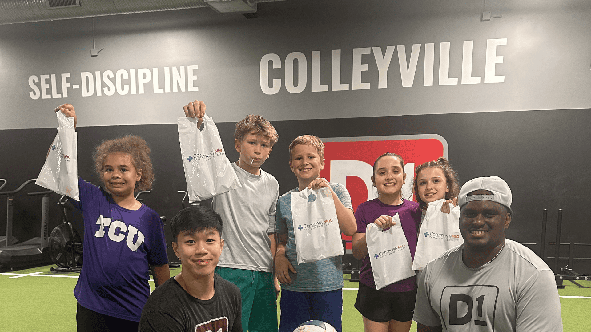 Group of six children holding CommunityMed snack bags alongside D1 coaches Kingsley and Lekieath in the Colleyville training turf